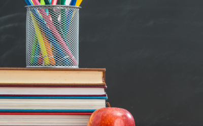 Colorful pens in a metal cup atop stacked books and a red apple, set against a chalkboard background, symbolizing back-to-school preparation and the start of the academic year.